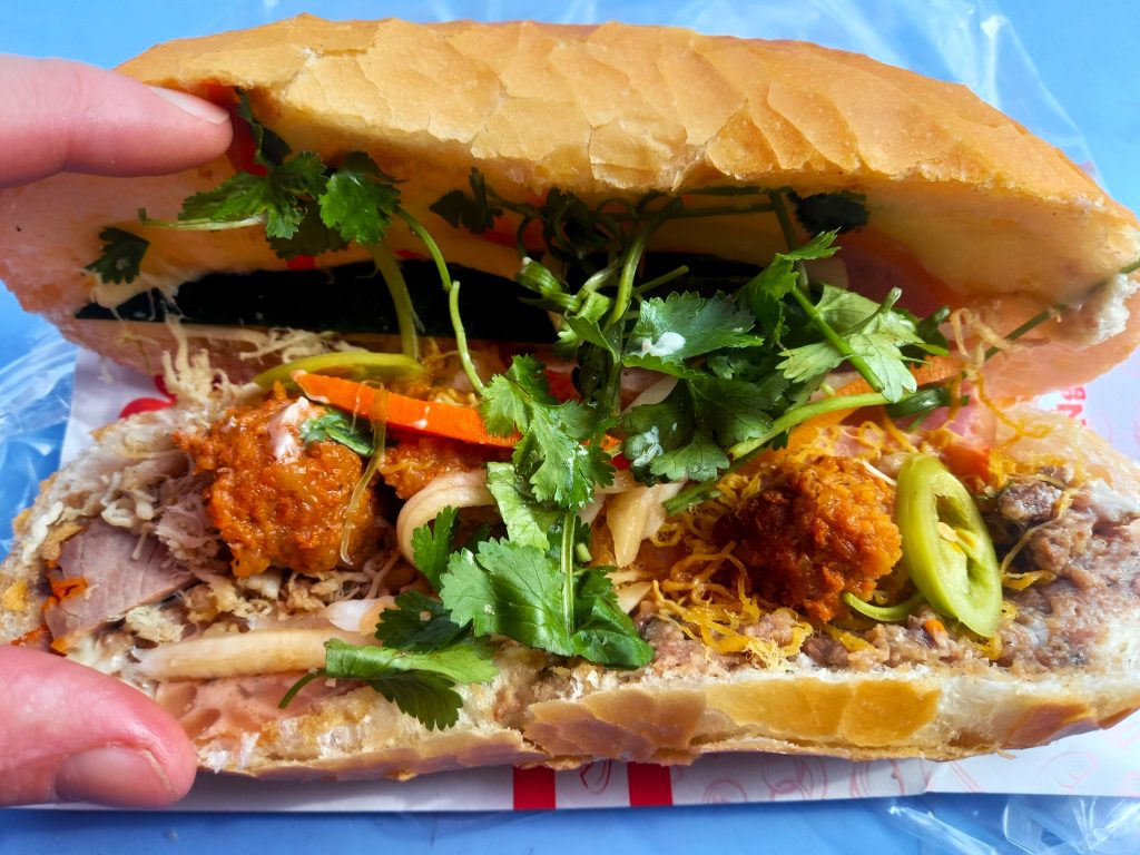 A Saigon-style banh mi sandwich held in hand, filled with pate, pork and fresh herbs
