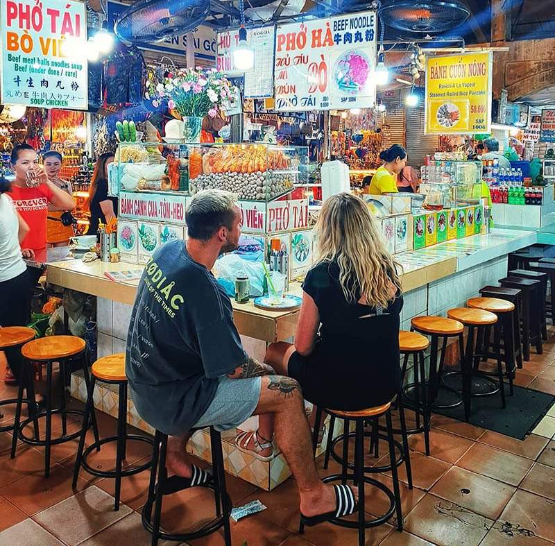 Two visitors on stools at a pho and noodle stall counter inside Ben Thanh Market