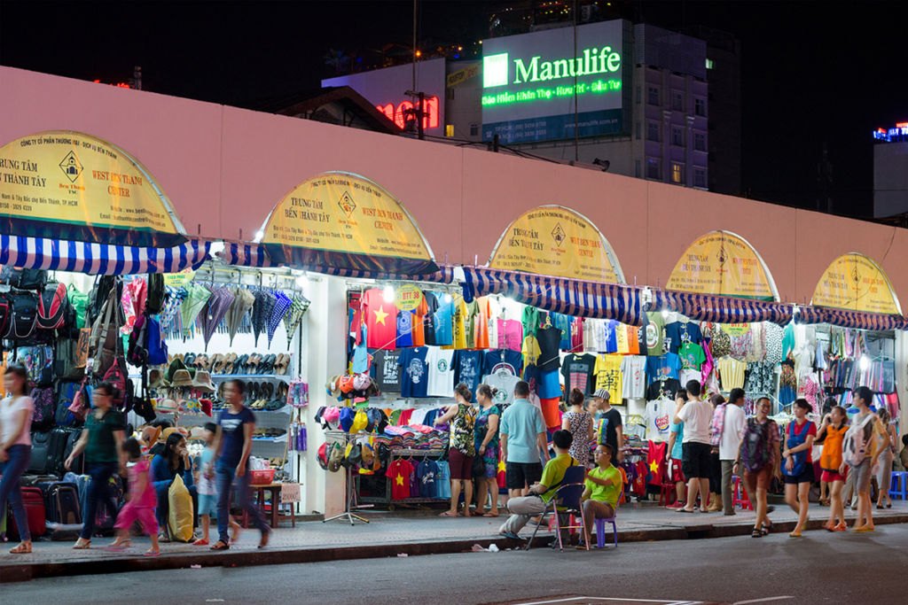 Clothes and souvenir stalls lit up along the outside wall of Ben Thanh Market at night