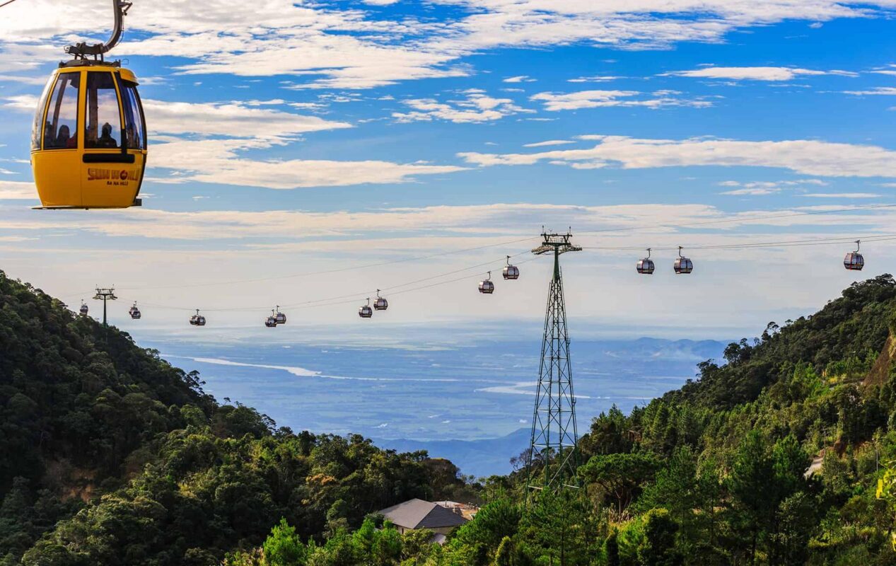 A line of cable car gondolas climbing over thick green forest towards a mountain