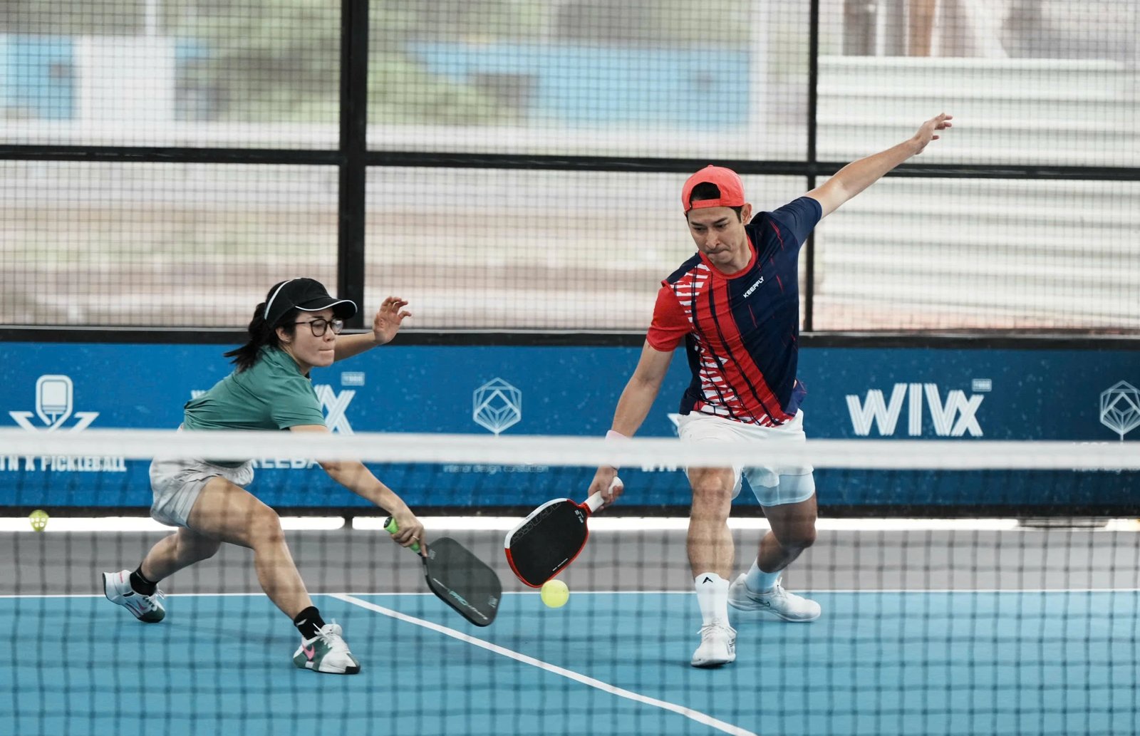 A mixed doubles pair stretching for a low ball at the net on a blue indoor court in Vietnam