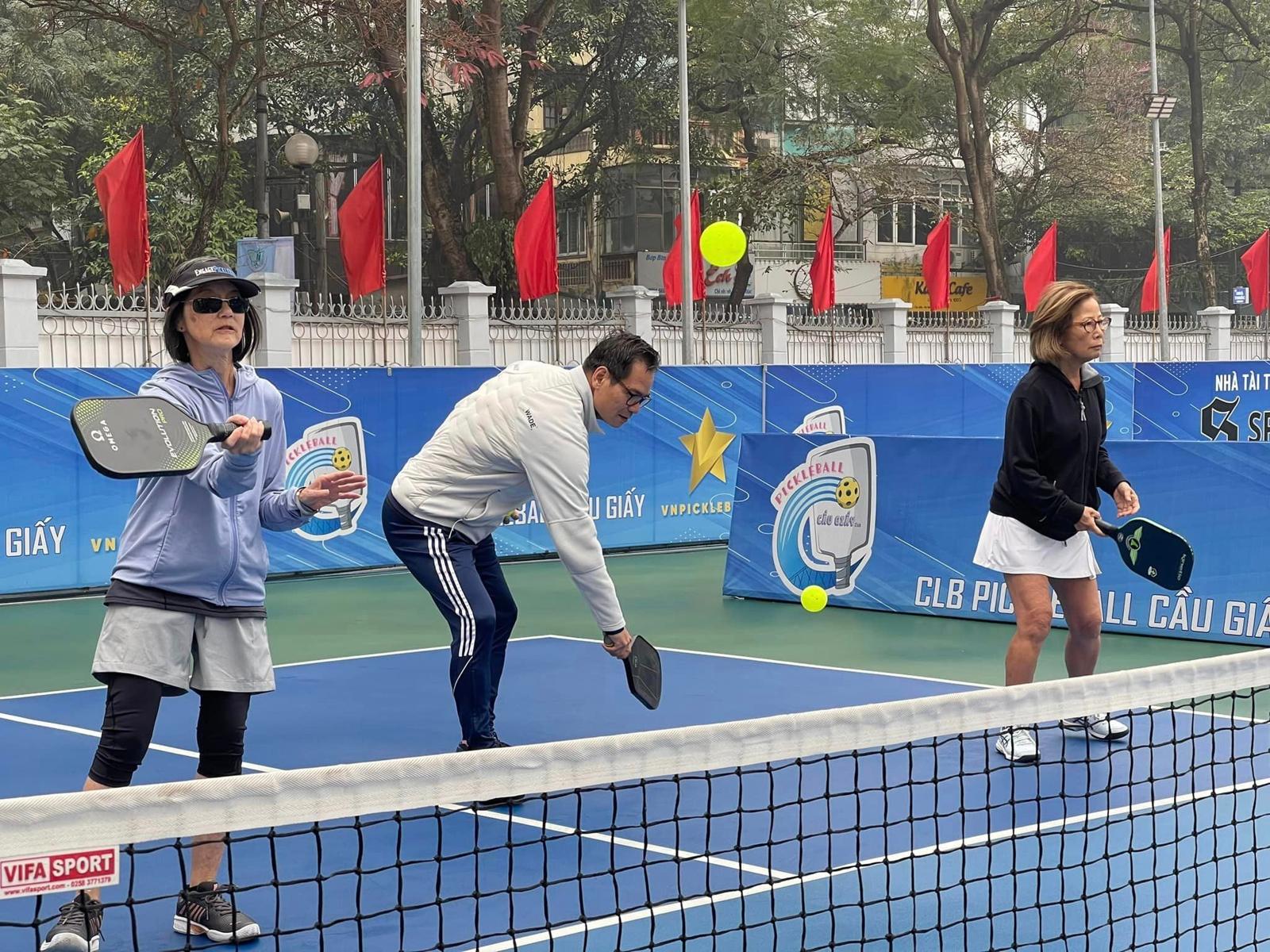 Three visiting players in jackets rallying at a doubles net on a blue outdoor court in Hanoi