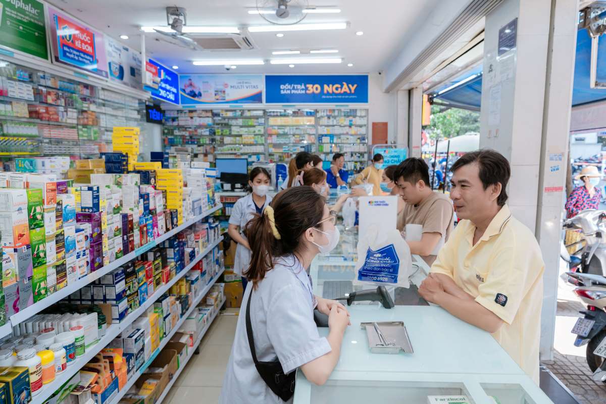 Customers and pharmacists inside a busy chain pharmacy in Vietnam, with shelves of medicines and pain relief