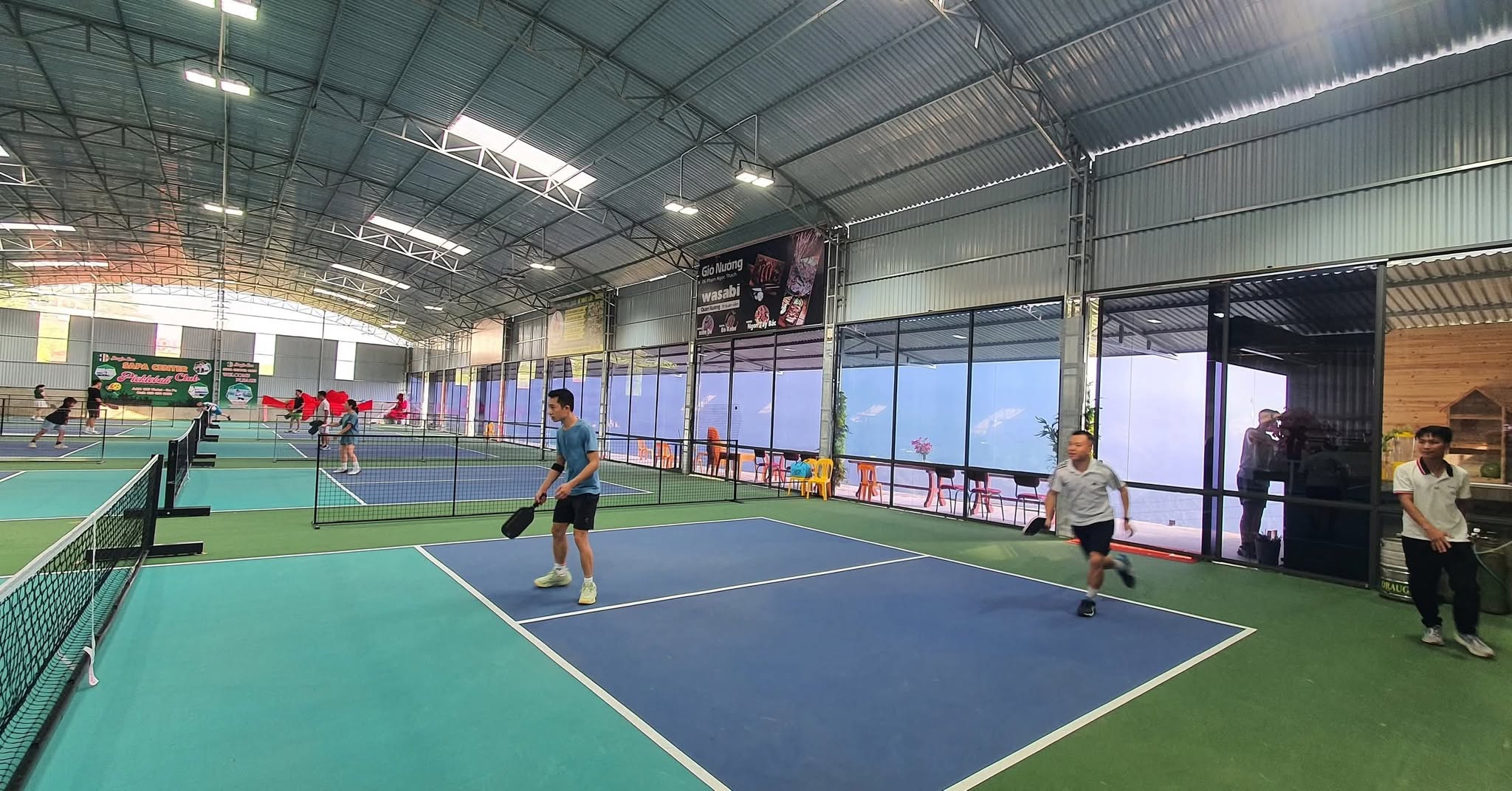 Players on blue and green courts inside a covered pickleball hall in Sapa with mountain views through the windows