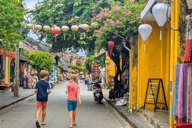 Two boys walking along a yellow-walled street in Hoi An old town under silk lanterns and bougainvillea